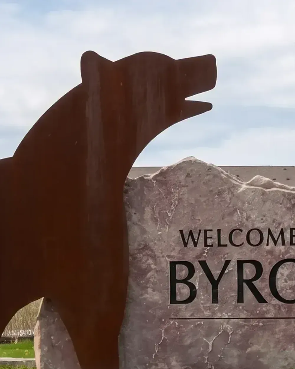The "Welcome to Byron" entrance sign in Byron, Minnesota, with a large rusted steel bear sculpture standing beside the carved stone marker.