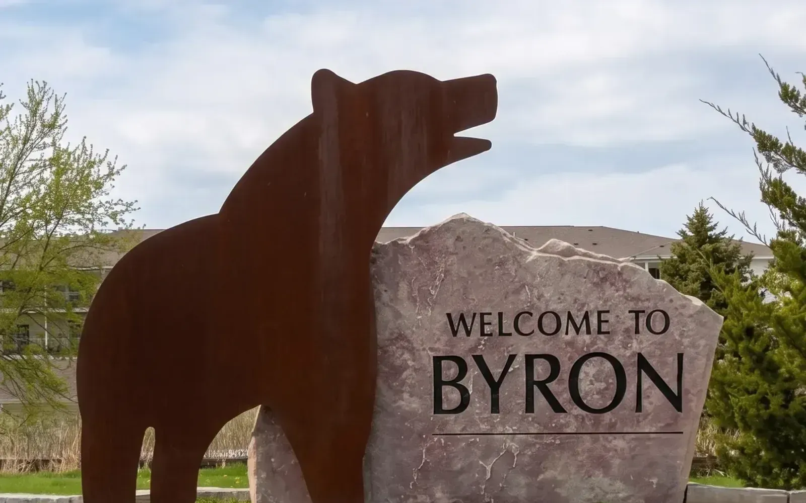 The "Welcome to Byron" entrance sign in Byron, Minnesota, with a large rusted steel bear sculpture standing beside the carved stone marker.