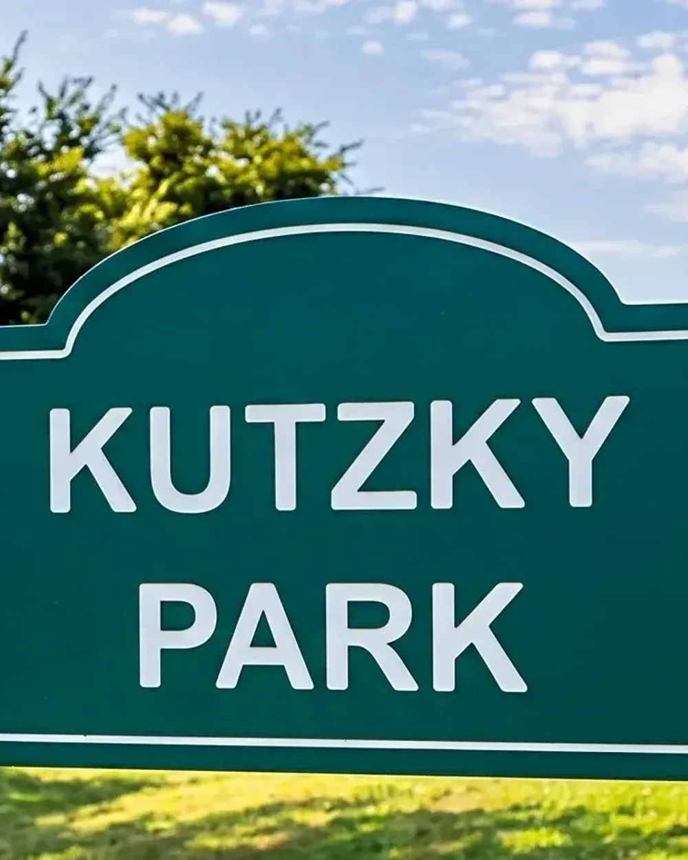 The Kutzky Park neighborhood sign in Rochester, Minnesota, set in the park lawn on a summer afternoon with mature trees in the background.