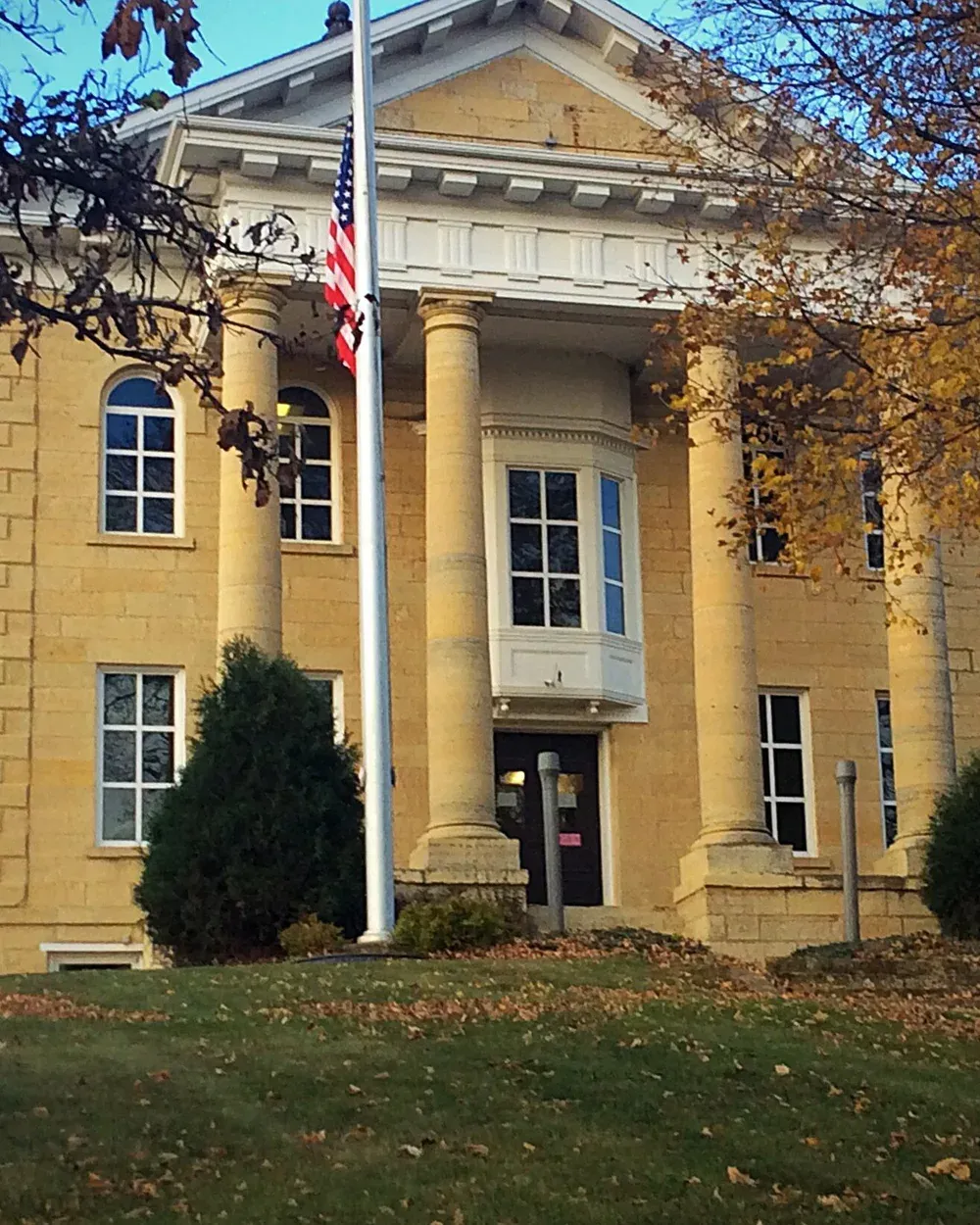 The 1865 Dodge County Courthouse in Mantorville, Minnesota, a Greek Revival limestone building with tall columns and a portico, surrounded by autumn foliage.