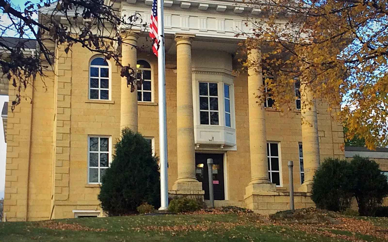 The 1865 Dodge County Courthouse in Mantorville, Minnesota, a Greek Revival limestone building with tall columns and a portico, surrounded by autumn foliage.